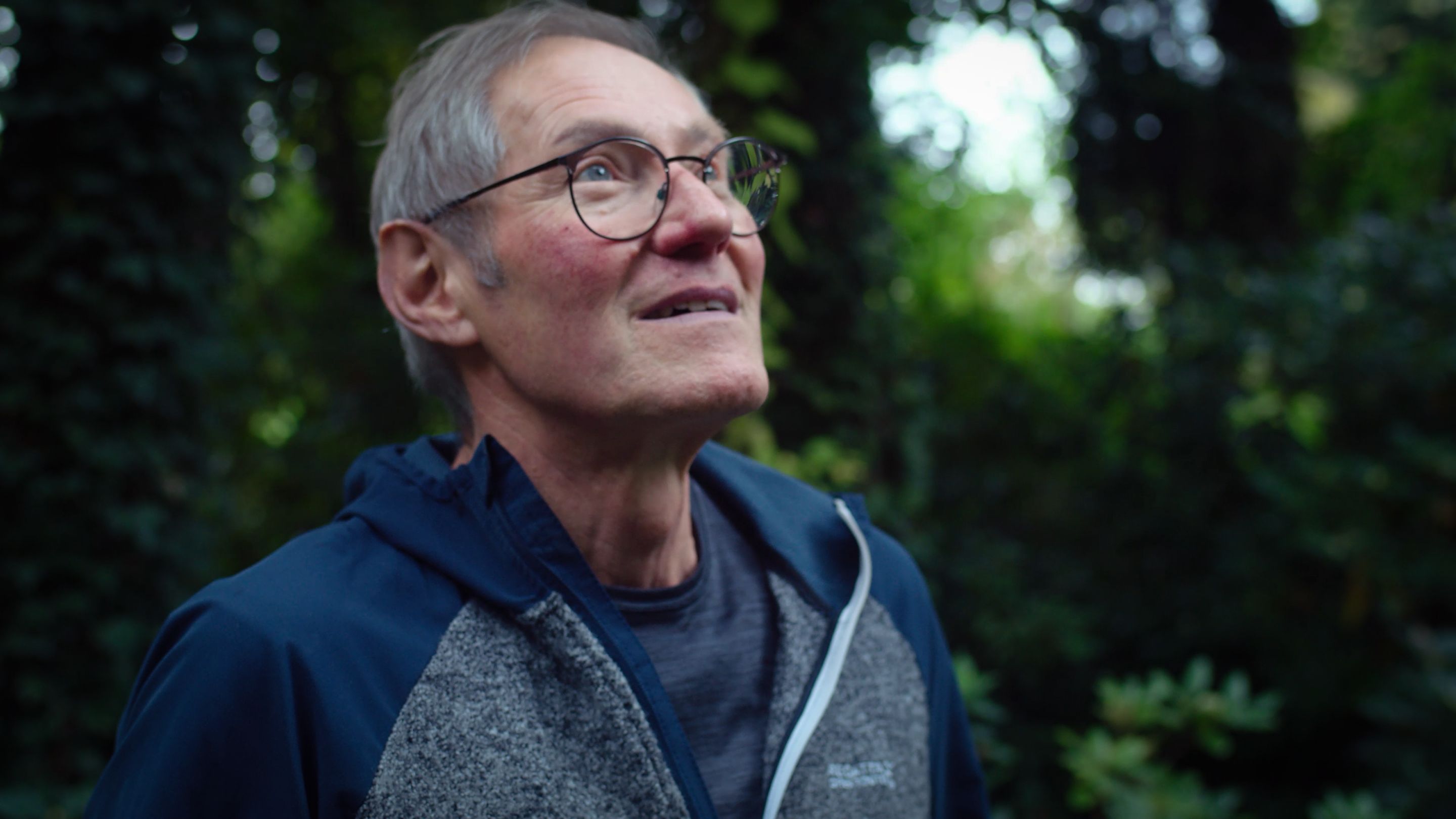 Older man with glasses looks upward outdoors, surrounded by greenery.