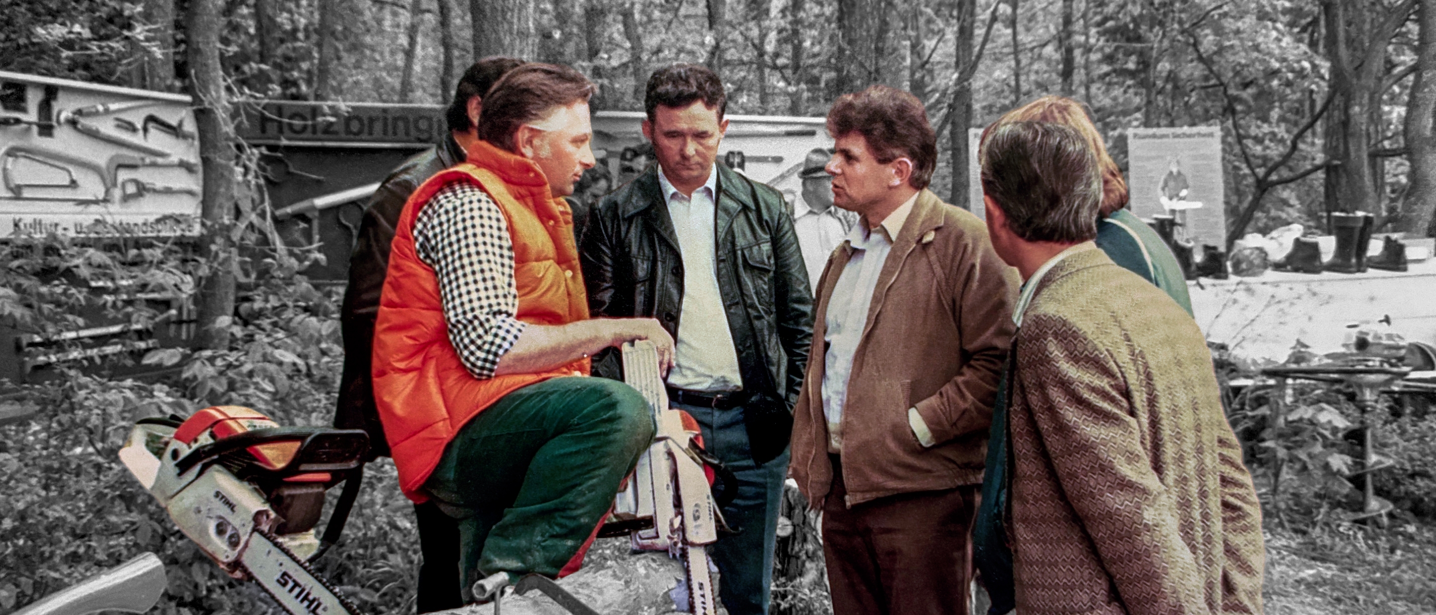 A group of men in the forest; one is wearing an orange vest.