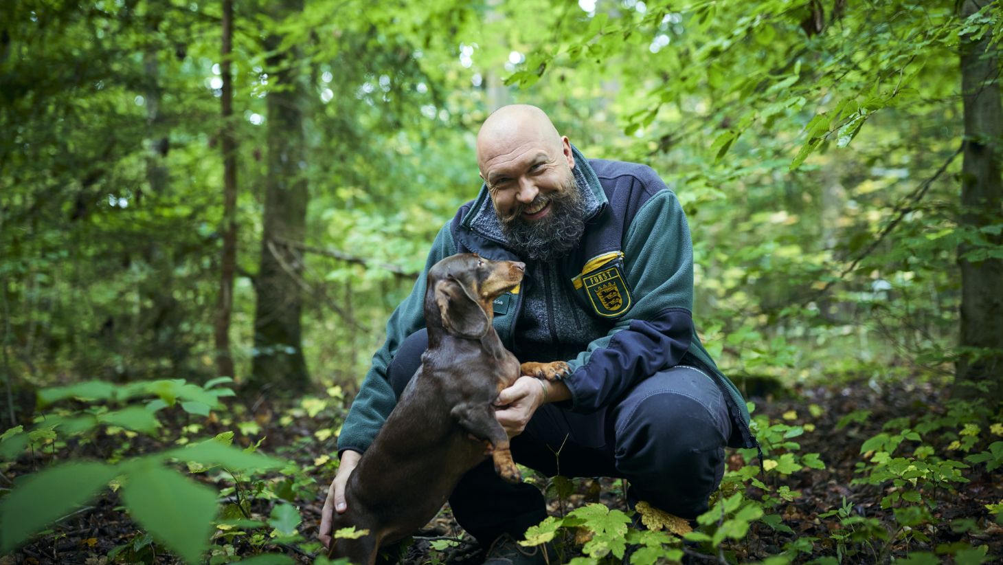 A man crouching in the forest with a dachshund.