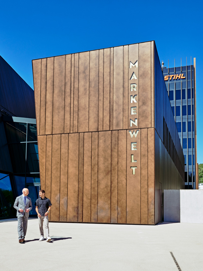 Two men walk to the left in front of a brown building with the &ldquo;Brand World&rdquo; sign.
