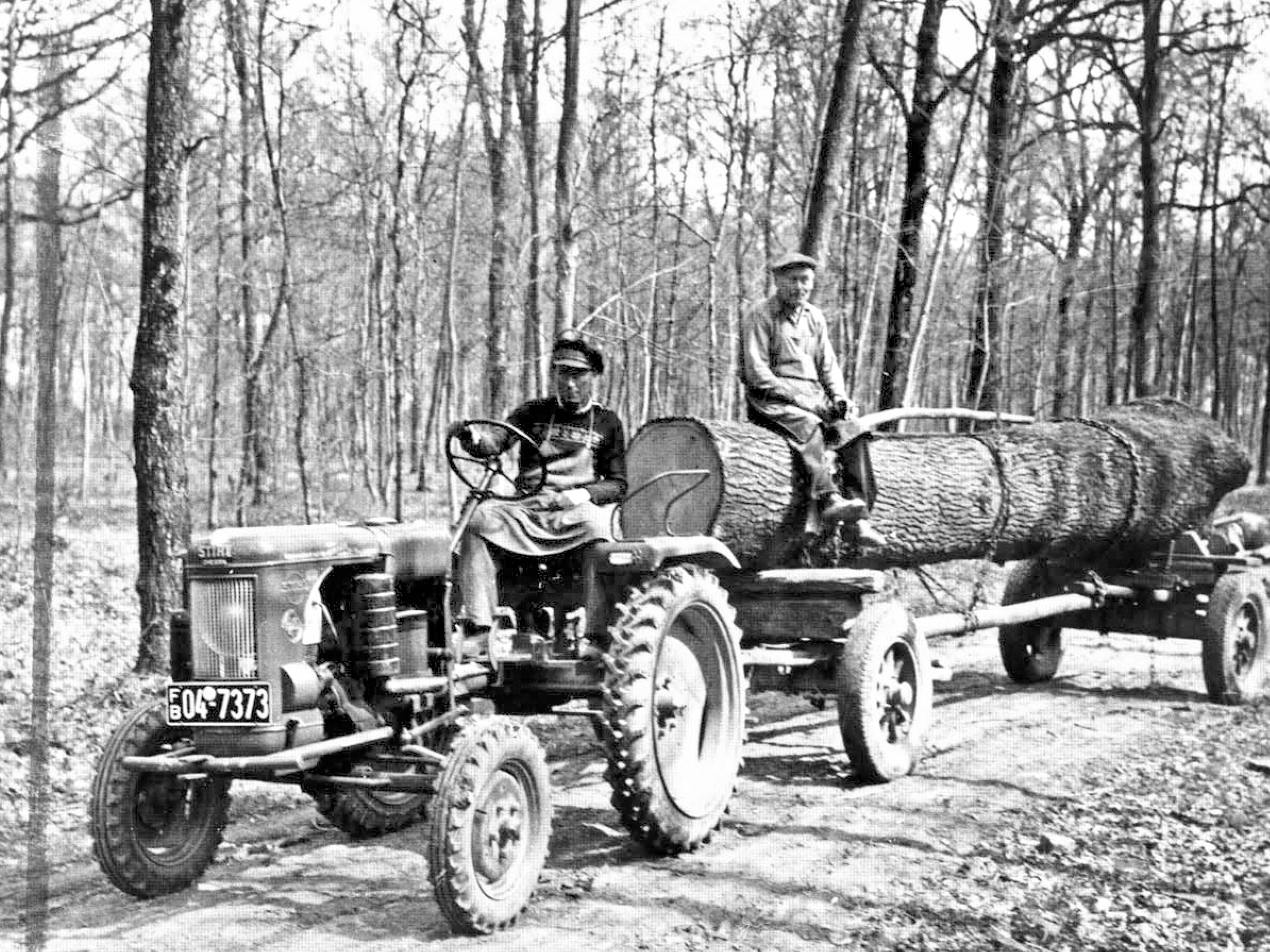 A man sits on a tractor and pulls a cart with a log; black-and-white photo.
