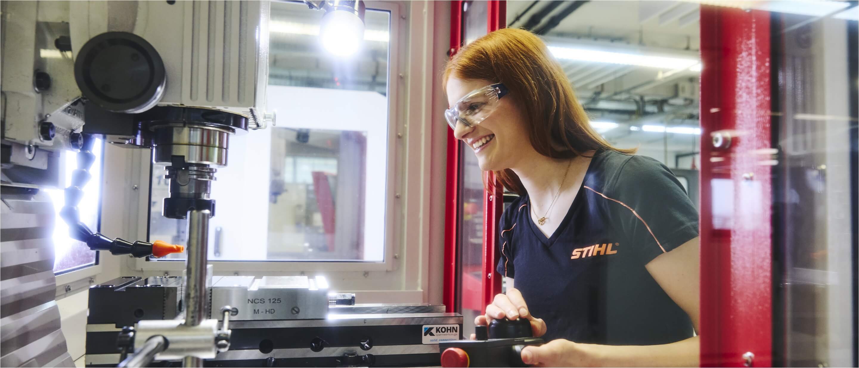 A young woman wearing safety glasses operating a milling machine with a remote control.