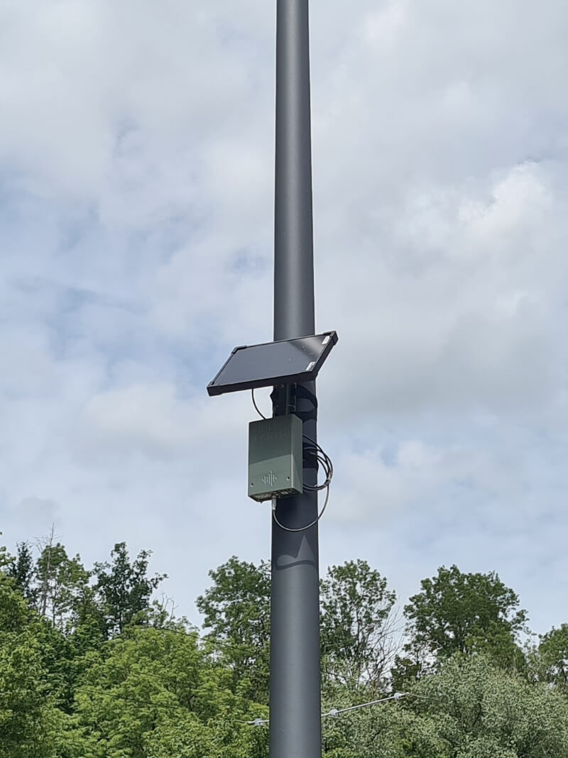 A lamppost with a rectangular box and a solar cell attached stands next to a brown building surrounded by trees.